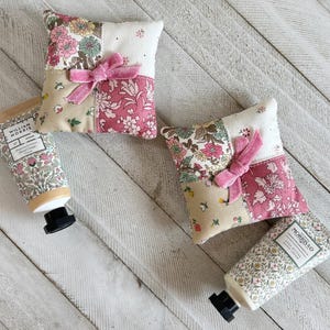 Puede incluir: Dos saquitos de tela cuadrados con patrones florales de patchwork en rosa, verde y crema, cada uno adornado con un pequeño lazo de terciopelo rosa. Dos tubos de crema de manos William Morris & Co. con diseños florales a juego están al lado.