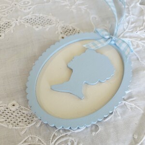 May include: A light blue wooden oval frame with a scalloped edge. The frame has a white background and a light blue silhouette of a person's head and shoulders inside.