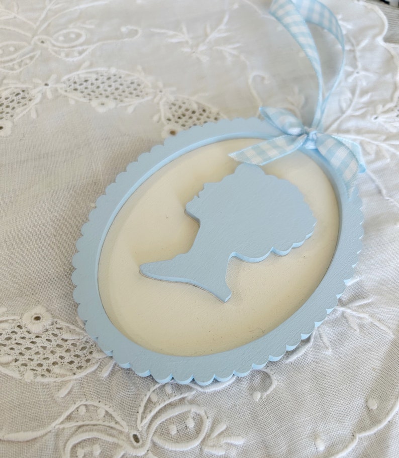 May include: A light blue wooden oval frame with a scalloped edge. The frame has a white background and a light blue silhouette of a person's head and shoulders inside.