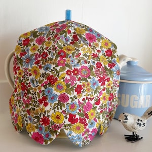 May include: A fabric tea cozy featuring a dense floral pattern with red, yellow, and blue flowers on a white background. The tea cozy has a scalloped bottom edge and a small blue fabric loop at the top center, covering a teapot.