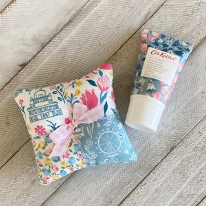 以下が含まれることがあります： ピンクのリボンボウが付いた小さな正方形のキルトのクッション。クッションには、青、ピンク、黄色の色合いのさまざまな花柄と幾何学模様が施されています。クッションの隣には、青と白の花柄の小さなハンドクリームのチューブがあります。チューブには「Cath Kidston Wild Barley & Meadows Hand Cream」とラベルが貼られています。