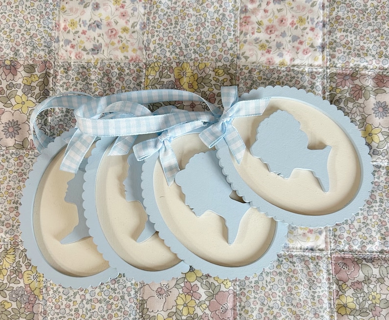 May include: Four light blue oval-shaped wooden frames with scalloped edges. Each frame features a white silhouette of a person's head and shoulders.