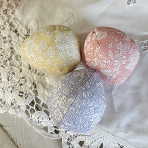 May include: Three fabric Easter eggs in pastel colors: yellow, pink, and lavender. Each egg is covered in a floral pattern with white flowers. The eggs are arranged on a white lace-trimmed surface.