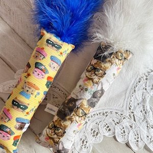 May include: Two cat toys with feather tops. One toy has a yellow background with cat faces wearing hats and masks. The other toy has a white background with cat faces wearing glasses. The toys have a rectangular shape.