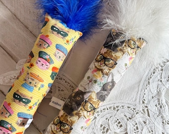 May include: Two cat toys with feather tops. One toy has a yellow background with cat faces wearing hats and masks. The other toy has a white background with cat faces wearing glasses. The toys have a rectangular shape.