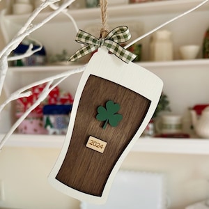 Puede incluir: Un adorno de madera blanco y marrón con forma de vaso de cerveza con un trébol verde y el año 2024 en él. El adorno está decorado con una cinta a cuadros verde y blanca.