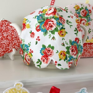 May include: A white teapot cozy with a floral pattern of red roses, blue and yellow flowers, and green leaves. The cozy has a scalloped edge and a red and white checkered loop at the top. Other tea cozies are visible in the background.