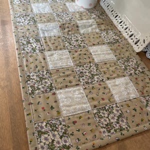 May include: A rectangular quilted table runner featuring a patchwork design of alternating squares. The fabric squares display a repeating pattern of small floral prints in shades of beige, white, pink, and green, finished with a matching fabric border.