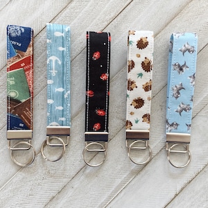 Può includere: Cinque portachiavi colorati con diversi motivi. I portachiavi sono realizzati in tessuto e hanno anelli metallici. I motivi includono un design di ancora blu e bianco, un design di coccinella nero e rosso, un design di riccio marrone e bianco, un design di gatto blu e bianco e un design a tema viaggio blu e bianco.