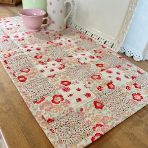 May include: A rectangular quilted table runner featuring a patchwork design of cream fabric squares with various floral patterns in shades of red, pink, and yellow. The quilted textile displays a repeating grid of alternating small and large floral prints.