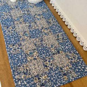 May include: A rectangular quilted table runner featuring a patchwork design of alternating blue and white fabric squares. The pattern displays floral motifs, small sailboats, and fish icons in shades of navy, light blue, and cream throughout.