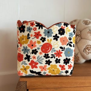 May include: A fabric storage bin with a scalloped top edge, featuring a cream-colored background printed with a repeating pattern of small flowers in shades of red, yellow, blue, and black, along with a central red circular face icon.