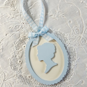 May include: A light blue oval ornament with a white background and a silhouette of a person's head and shoulders. The ornament has a blue and white gingham ribbon attached.