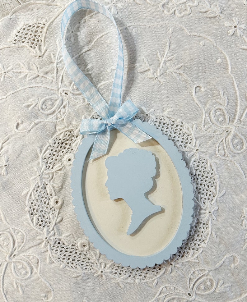 May include: A light blue oval ornament with a white background and a silhouette of a person's head and shoulders. The ornament has a blue and white gingham ribbon attached.