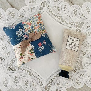 以下が含まれることがあります： 茶色のリボンが付いた小さなパッチワーク枕と、ウィリアム・モリスのストロベリーシーフハンドクリームのチューブ。枕は青、ピンク、白の花柄模様です。ハンドクリームチューブはベージュの花柄模様と黒いキャップが付いています。