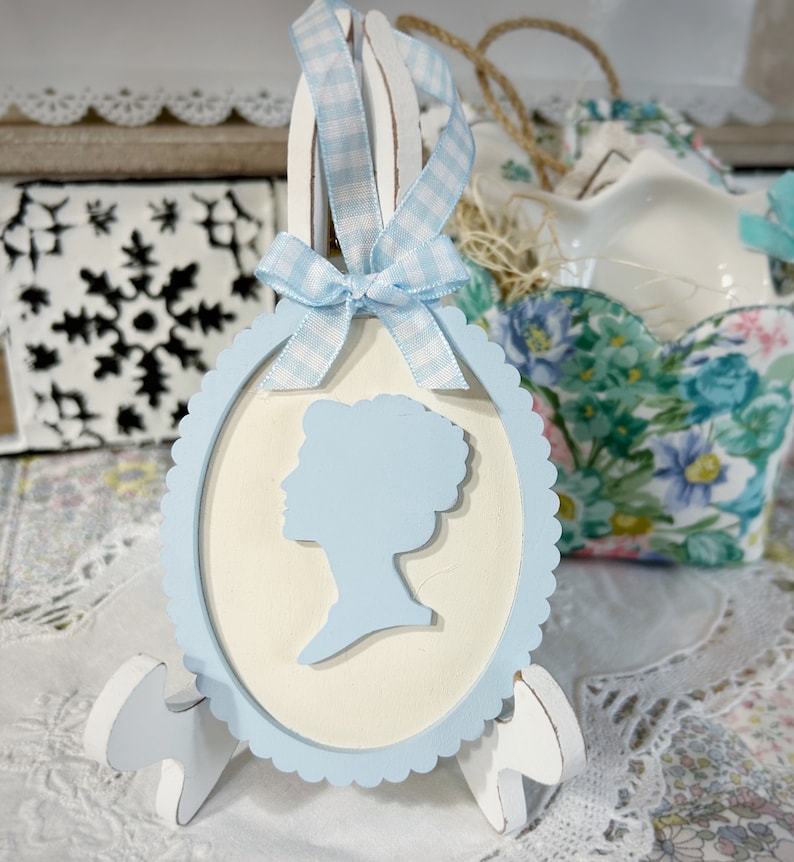 May include: A light blue and white oval wooden ornament with a cutout silhouette of a woman's head. The ornament is decorated with a blue and white gingham ribbon bow and is displayed on a white wooden stand.