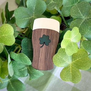 May include: Wooden beer glass cutout with a green shamrock design. The top of the glass is white, and the body is brown with wood grain. The cutout is surrounded by green shamrock leaves.