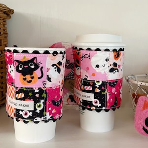 May include: Two white coffee cups featuring fabric sleeves with a patchwork Halloween print in pink, black, and white. The sleeves display ghosts, black cats, pumpkins, and candy, with a black scalloped trim and a white label reading elliebug makes.