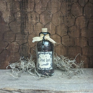May include: A small glass bottle with a black wax seal and a label that reads "CC 18 Dried Leech". The bottle is filled with dried black leeches and is sitting on a wooden surface.