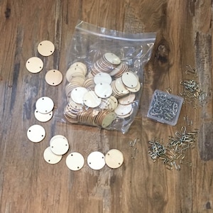 May include: A collection of small, round, unfinished wooden discs with pre-drilled holes, scattered on a wooden surface. A clear plastic bag contains a larger quantity of the same discs. Also included are small metal fasteners.