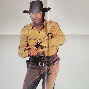 Peut inclure: Un homme portant un chapeau de cowboy marron et une chemise jaune tient une arme à feu. Il porte un pantalon marron et une ceinture marron avec une boucle argentée. Il se tient devant un fond blanc.
