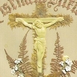 May include: A vintage religious image featuring a white ceramic figure of Jesus Christ on the cross. The figure is surrounded by dried ferns and flowers, with the text "In Mein Herz" embroidered in brown thread on the background.