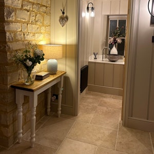May include: A white console table with turned legs sits in a hallway with a stone wall and a doorway leading to a bathroom. The table has a wooden top and a lamp with a white and blue patterned shade. A vase with white flowers sits on the table.
