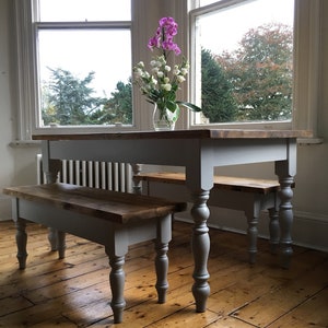 May include: A grey farmhouse style dining table with two matching benches. The table and benches have turned legs and a natural wood top. The table is set in a room with hardwood floors and large windows.