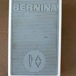 May include: A gray and white Bernina sewing machine foot pedal. The pedal has a textured surface with the brand name "BERNINA" in capital letters. A circular emblem with a needle and diamond shapes is also visible.
