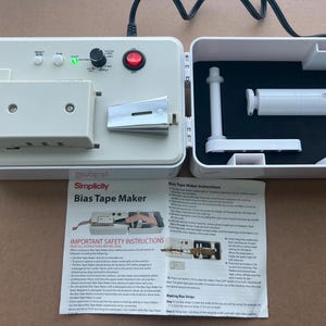 Op de afbeelding: Een witte Simplicity Bias Tape Maker met een zwart netsnoer. De machine heeft een bedieningspaneel met knoppen en een draaiknop. De open behuizing onthult witte accessoires op een zwart oppervlak. Een gedrukte instructievel ligt ervoor.