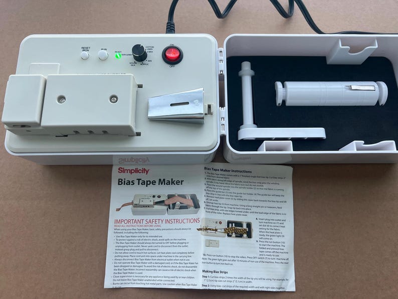 Op de afbeelding: Een witte Simplicity Bias Tape Maker met een zwart netsnoer. De machine heeft een bedieningspaneel met knoppen en een draaiknop. De open behuizing onthult witte accessoires op een zwart oppervlak. Een gedrukte instructievel ligt ervoor.