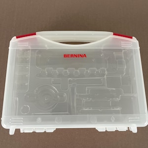 May include: A clear plastic storage case with a handle and red accents, featuring the word BERNINA printed in red on the front. The interior is molded with various compartments for organizing sewing machine accessories and tools.