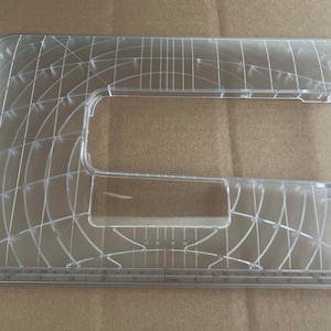 May include: Clear plastic quilting template with a grid pattern and a ruler along the bottom edge. The template has a rectangular shape with rounded corners and a cutout in the center. Measurements are in inches.