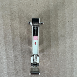 May include: A silver-colored metal sewing machine zipper foot attachment featuring a vertical rectangular white label with a red number 4 and a small pink and white striped pattern at the top, designed with a hinged base and a top mounting bar.