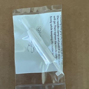May include: A white plastic needle insertion tool packaged in a clear plastic bag with an instruction card. The card features a line drawing of a hand and text that reads: Holding the grip (A), insert the needle into the needle clamp.
