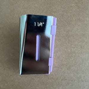 May include: A silver-colored bias tape maker with a 1 1/4" measurement printed on the front. The tool has a purple guide and is designed for creating bias tape for sewing projects.