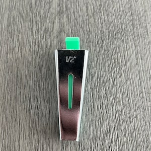 May include: A silver-colored bias tape maker with a green plastic guide. The tool is marked with "1/2" and is designed to fold fabric strips into bias tape. The tool is used for sewing and crafting projects.