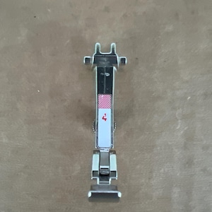 May include: A silver-colored metal sewing machine presser foot attachment featuring a vertical rectangular white label with a red diagonal striped pattern at the top and the red number 4 printed clearly in the center of the white section.