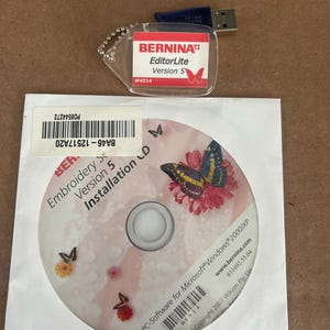 May include: A Bernina EditorLite Version 5 USB drive and CD-ROM for embroidery software. The CD-ROM is in a white paper sleeve and features butterfly and flower graphics. The USB drive is attached to a clear plastic tag.