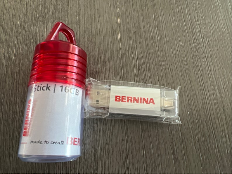 May include: A white USB stick with the word "BERNINA" printed on it. The stick is in a clear plastic case. The stick is next to a red plastic case with the words "Stick | 16GB" and "made to create" printed on it.