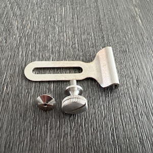 May include: A silver metal bracket with two screws. The bracket has a slot in the middle and a curved end. The screws are silver and have a knurled head.