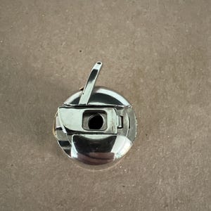 May include: A silver-colored metal sewing machine bobbin case with a circular body, a hinged latch, and a small upright tension arm. The component features a central opening and a polished metallic finish, designed for holding a bobbin in a machine.