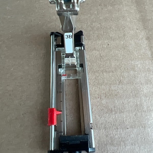 May include: A silver sewing machine presser foot with a black base and a red adjustment lever. The presser foot has the number "3B" printed on a white label. The item is designed for sewing applications.