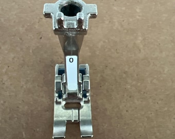 Op de afbeelding: Een zilverkleurige naaivoet voor naaimachines, met een rechthoekige basis en een cilindrische bovenkant. Het nummer "0" is gedrukt op een wit label. Ontworpen voor naaimachines.