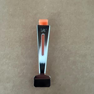 May include: A silver-colored sewing machine attachment with an orange top and a black base. The text "1/4" is printed on the attachment. The tool is designed for sewing and crafting projects.