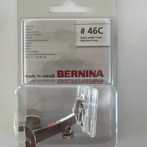 Op de afbeelding: Een zilveren naaimachinevoet in een doorzichtige plastic verpakking. De voet is gemarkeerd met #46C, steekbreedte 9 mm, machine 9 mm. De verpakking vermeldt ook "made to create BERNINA".