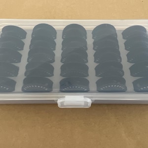 May include: A clear plastic storage box with a white latch. The box is filled with 36 black guitar picks.