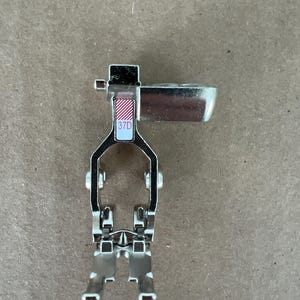 May include: A silver-colored metal sewing machine presser foot attachment featuring a dual-prong design at the base. The component includes a small rectangular label with red diagonal stripes and the alphanumeric text 37D printed in red ink.