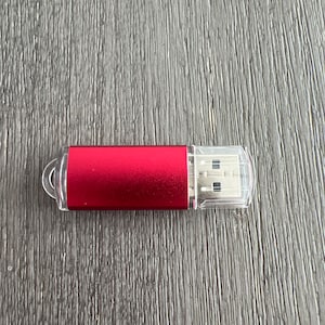 Op de afbeelding: Een rode USB-stick met een doorzichtige plastic dop. De USB-stick is rechthoekig met afgeronde randen en een kleine lus om aan een sleutelhanger te bevestigen. De stick ligt op een grijs houten oppervlak.