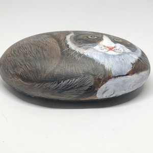 May include: Hand-painted rock art depicting a gray and white cat with green eyes. The cat is painted in a realistic style and is lying down on its side.